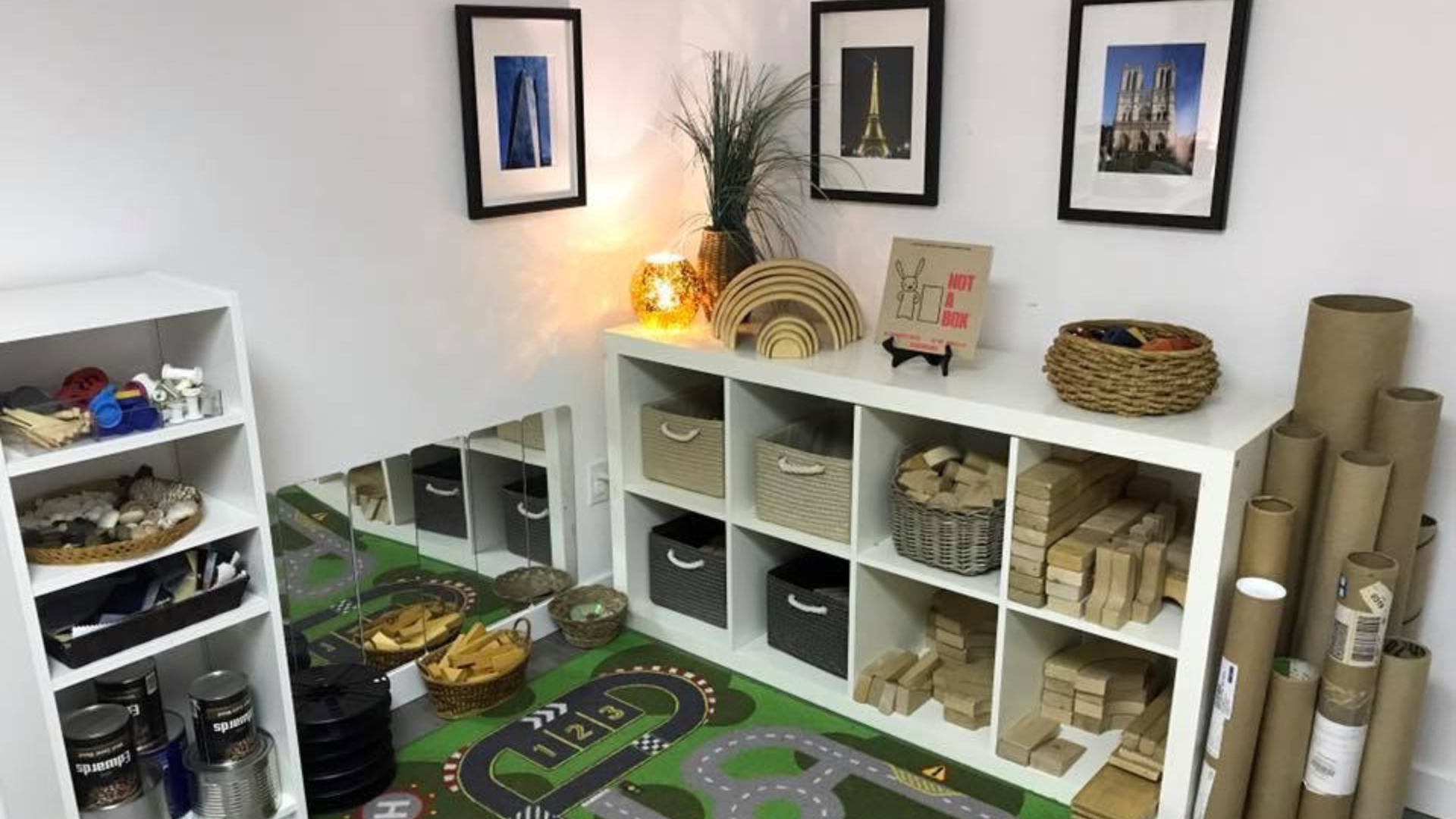 A well-organized early childhood learning environment featuring shelves with baskets of wooden blocks, cardboard tubes, and various loose parts. A soft lamp illuminates the corner, which is adorned with framed pictures and natural elements.